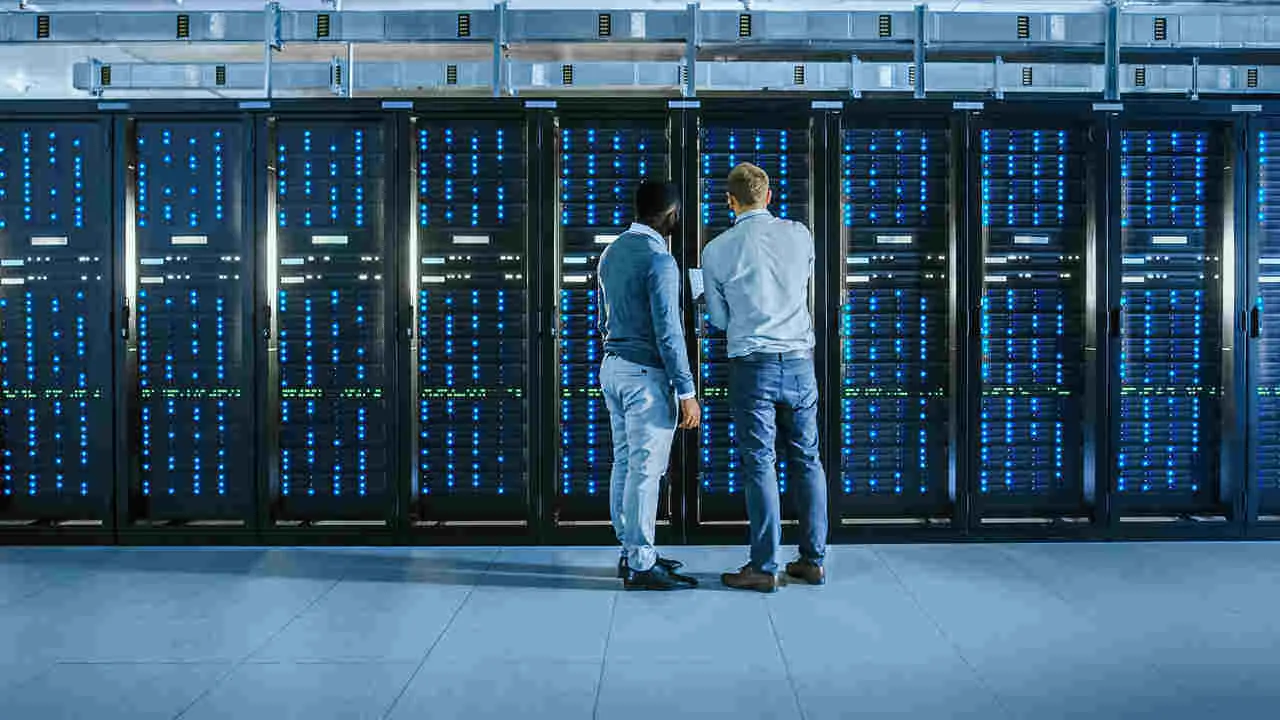 IT technician in glasses with a laptop and male engineer colleague are talking in data center while working next to server racks