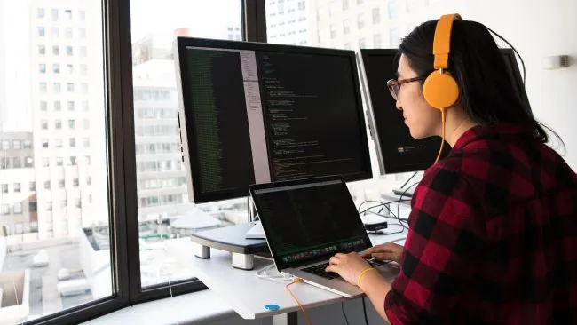 woman with orange headphones working on laptop and bigger screen at her desk in front of a window 